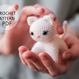 May include: A small, crocheted white cat with blue eyes and a pink nose is held in cupped hands. The cat has small, pointed ears and a curled tail. The text "CROCHET PATTERN PDF" is in a white circle in the upper left corner.