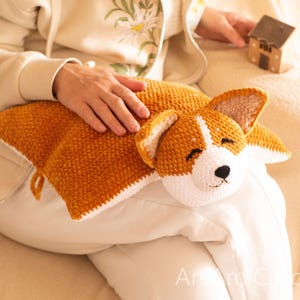 Peut inclure: Un coussin rectangulaire en crochet repr&eacute;sentant un chien Corgi. Le coussin pr&eacute;sente un corps textur&eacute; brun dor&eacute; avec un dessous blanc et une t&ecirc;te de Corgi d&eacute;taill&eacute;e avec des oreilles pointues, des yeux ferm&eacute;s et un nez noir. Le texte AniCrochet est visible.