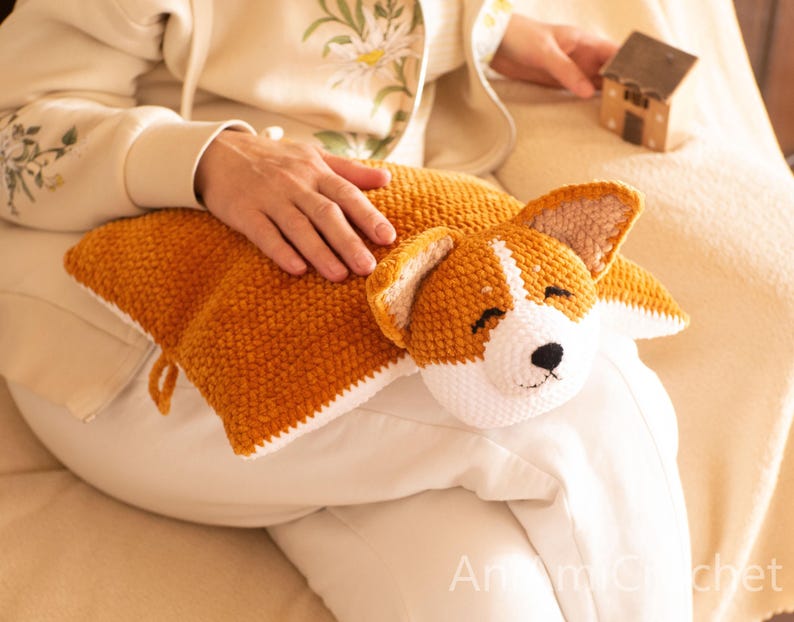 Peut inclure: Un coussin rectangulaire en crochet repr&eacute;sentant un chien Corgi. Le coussin pr&eacute;sente un corps textur&eacute; brun dor&eacute; avec un dessous blanc et une t&ecirc;te de Corgi d&eacute;taill&eacute;e avec des oreilles pointues, des yeux ferm&eacute;s et un nez noir. Le texte AniCrochet est visible.