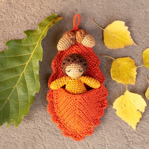 May include: A crocheted acorn-shaped pouch with a brown and yellow crocheted doll inside. The pouch is orange and has a scalloped edge. The doll is lying down with its arms outstretched. The pouch is surrounded by yellow leaves.