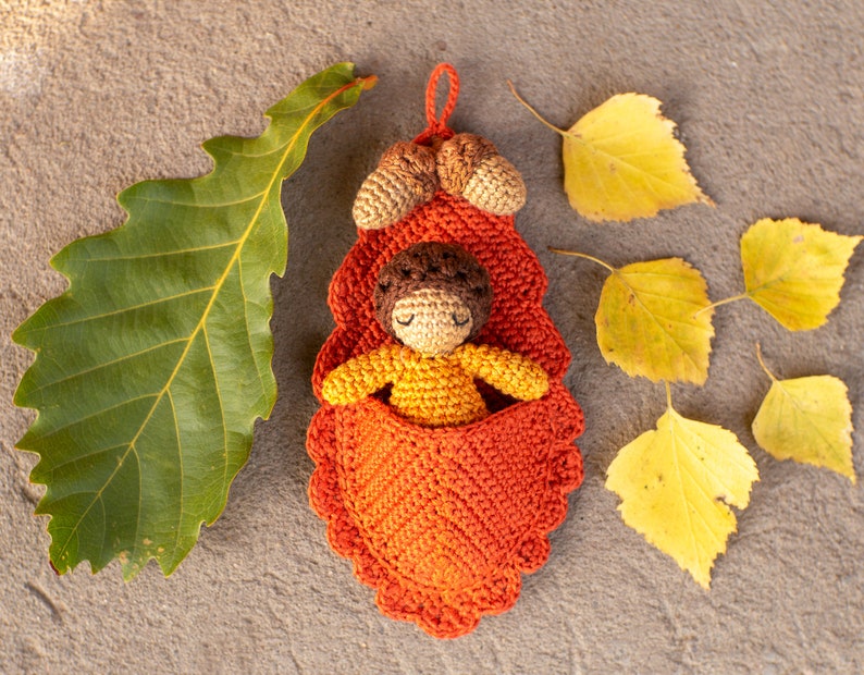 May include: A crocheted acorn-shaped pouch with a brown and yellow crocheted doll inside. The pouch is orange and has a scalloped edge. The doll is lying down with its arms outstretched. The pouch is surrounded by yellow leaves.