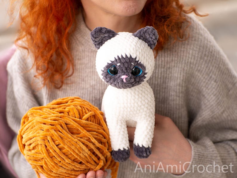 K&ouml;nnte beinhalten: Ein handgefertigtes H&auml;kel-Pl&uuml;schtier in Form einer Siamkatze aus wei&szlig;em und dunkelgrauem Garn mit gro&szlig;en blauen Augen und einer kleinen rosa gestickten Nase. Neben der Pl&uuml;schkatze liegt ein gro&szlig;es Kn&auml;uel goldgelbes Samtgarn. Text AniAmiCrochet.