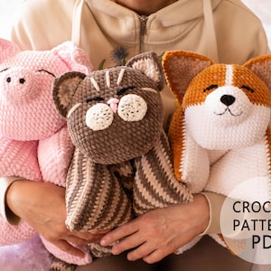 May include: Three crocheted soft toys: a pink pig, a brown cat, and an orange and white corgi. The pig has a smiling face, the cat has striped details, and the corgi has large ears. The image includes the text "CROCHET PATTERN PDF".