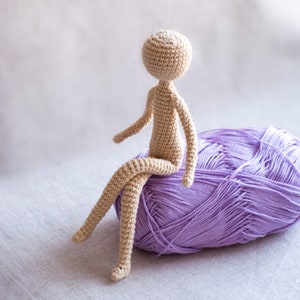 K&ouml;nnte beinhalten: Eine geh&auml;kelte Puppe mit einem beigefarbenen K&ouml;rper und einem hellvioletten Garnkn&auml;uel.