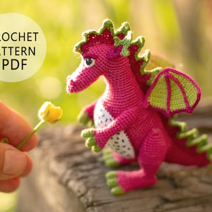 Puede incluir: Un dragón de crochet rosa con detalles verdes y manchas blancas. El dragón está de pie sobre una superficie de madera y tiene las alas extendidas. Una mano sostiene una flor amarilla frente al dragón.