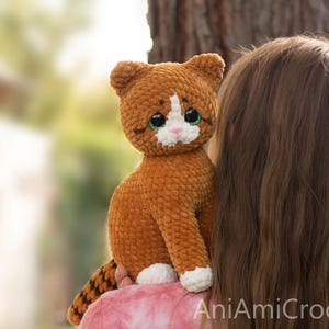 May include: A handmade crochet cat plush toy featuring soft, textured brown yarn, a white patch on its face, white paws, and large green eyes. The cat has a striped tail and a small pink nose. The text AniAmiCrochet is visible in the bottom corner.