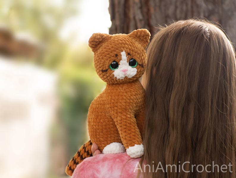 May include: A handmade crochet cat plush toy featuring soft, textured brown yarn, a white patch on its face, white paws, and large green eyes. The cat has a striped tail and a small pink nose. The text AniAmiCrochet is visible in the bottom corner.