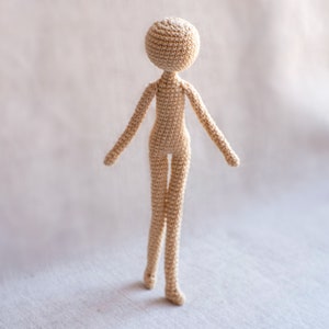 K&ouml;nnte beinhalten: Eine geh&auml;kelte Puppe mit einem beigefarbenen K&ouml;rper und einem runden Kopf. Die Puppe steht auf einer wei&szlig;en Oberfl&auml;che.