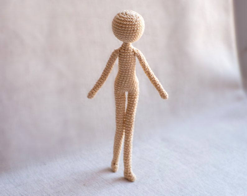 K&ouml;nnte beinhalten: Eine geh&auml;kelte Puppe mit einem beigefarbenen K&ouml;rper und einem runden Kopf. Die Puppe steht auf einer wei&szlig;en Oberfl&auml;che.