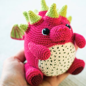 Op de afbeelding: Een gehaakt draken knuffel met een roze lichaam, groene stekels en witte vlekken. De draak heeft zwarte ogen en een witte mond.