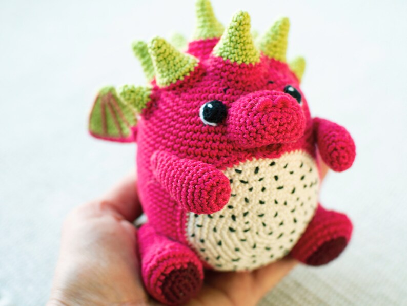 Op de afbeelding: Een gehaakt draken knuffel met een roze lichaam, groene stekels en witte vlekken. De draak heeft zwarte ogen en een witte mond.