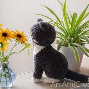 May include: A handmade crochet cat plush toy with dark gray and white yarn, featuring a rounded head, pointed ears, and a long tail. The cat is positioned in profile, facing a clear glass vase filled with yellow flowers with dark brown centers.