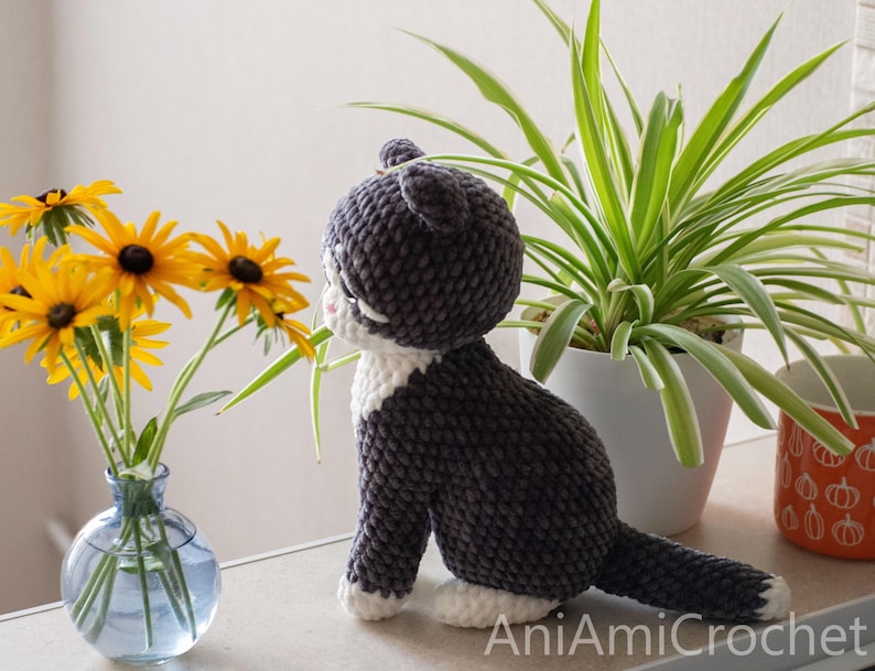 May include: A handmade crochet cat plush toy with dark gray and white yarn, featuring a rounded head, pointed ears, and a long tail. The cat is positioned in profile, facing a clear glass vase filled with yellow flowers with dark brown centers.
