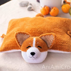 Peut inclure: Un coussin rectangulaire en crochet en forme de corgi, avec un corps brun dor&eacute;, un dessous blanc et une petite queue. La t&ecirc;te attach&eacute;e pr&eacute;sente des oreilles pointues, un museau blanc, un nez noir et des yeux ferm&eacute;s. Le texte AniAmiCrochet est visible.