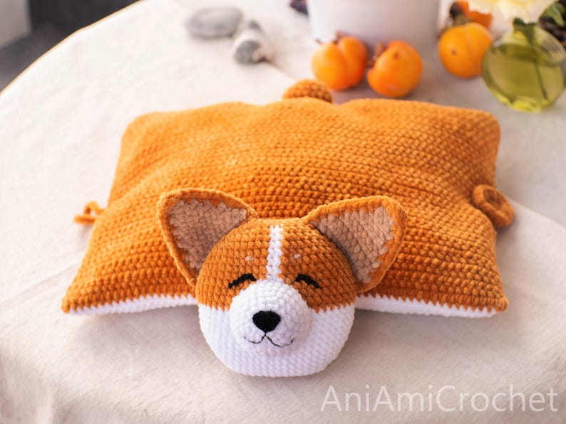 Peut inclure: Un coussin rectangulaire en crochet en forme de corgi, avec un corps brun dor&eacute;, un dessous blanc et une petite queue. La t&ecirc;te attach&eacute;e pr&eacute;sente des oreilles pointues, un museau blanc, un nez noir et des yeux ferm&eacute;s. Le texte AniAmiCrochet est visible.