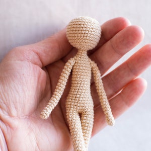 K&ouml;nnte beinhalten: Eine kleine, geh&auml;kelte Puppe in beige. Die Puppe hat einen runden Kopf, Arme und Beine. Die Puppe wird in der Hand einer Person gehalten.