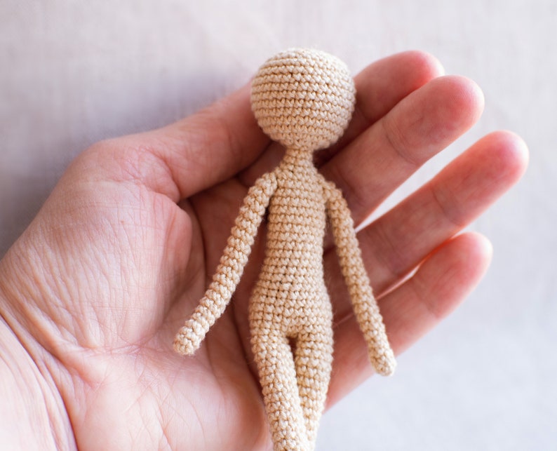 K&ouml;nnte beinhalten: Eine kleine, geh&auml;kelte Puppe in beige. Die Puppe hat einen runden Kopf, Arme und Beine. Die Puppe wird in der Hand einer Person gehalten.