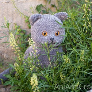 May include: A gray crocheted cat with orange eyes sits amidst green foliage and yellow flowers. The handmade plush toy has a detailed, textured appearance, with visible stitches and a soft, velvety texture. The cat's ears are perked up, and it appears to be looking forward.