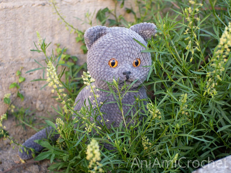 May include: A gray crocheted cat with orange eyes sits amidst green foliage and yellow flowers. The handmade plush toy has a detailed, textured appearance, with visible stitches and a soft, velvety texture. The cat's ears are perked up, and it appears to be looking forward.