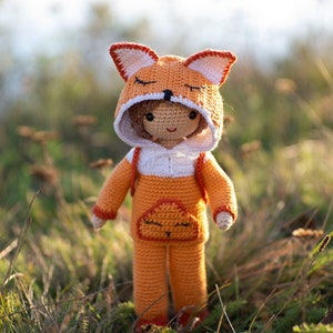 May include: A crocheted fox doll wearing an orange and white hooded jumpsuit. The doll has a sweet expression and is standing in a field of tall grass.