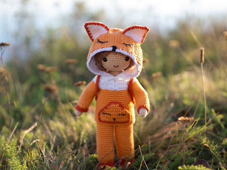 May include: A crocheted fox doll wearing an orange and white hooded jumpsuit. The doll has a sweet expression and is standing in a field of tall grass.