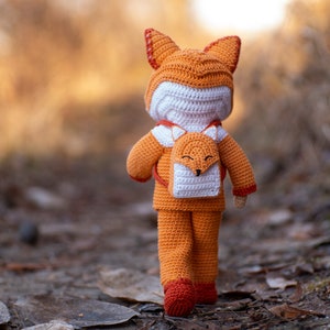 May include: A crocheted fox doll wearing an orange and white fox-shaped backpack. The doll is standing on a path in a forest.
