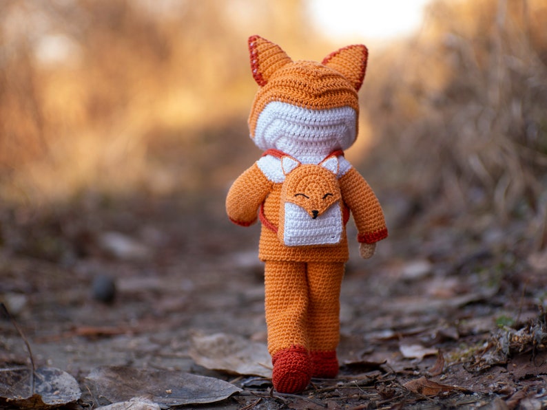 May include: A crocheted fox doll wearing an orange and white fox-shaped backpack. The doll is standing on a path in a forest.