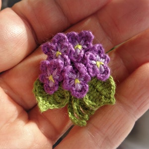 May include: A small crocheted bouquet of purple flowers with yellow centers and green leaves.