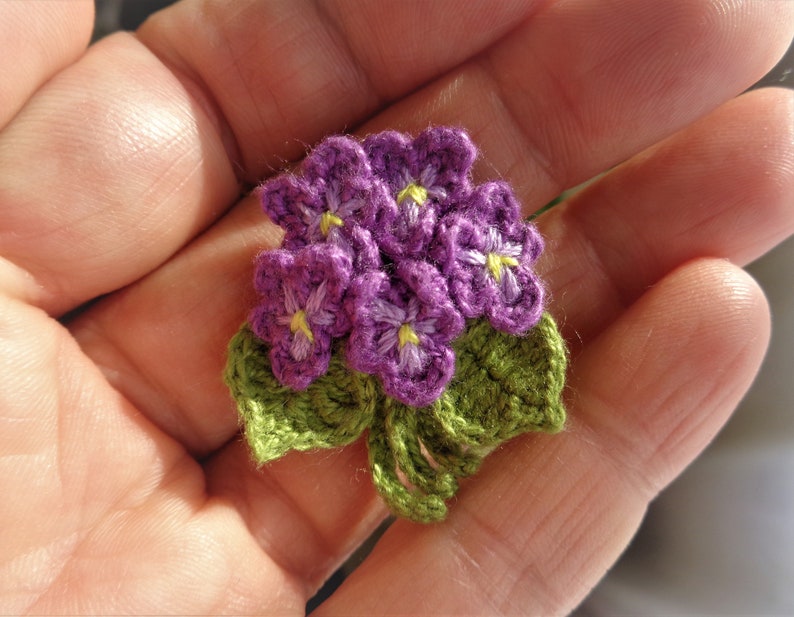 May include: A small crocheted bouquet of purple flowers with yellow centers and green leaves.
