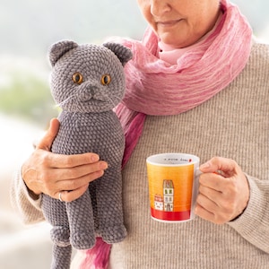 May include: A gray crocheted cat with orange eyes and a white whisker is held next to a mug. The mug has a yellow and orange gradient with a red base and features a house illustration. The cat is a plush toy.