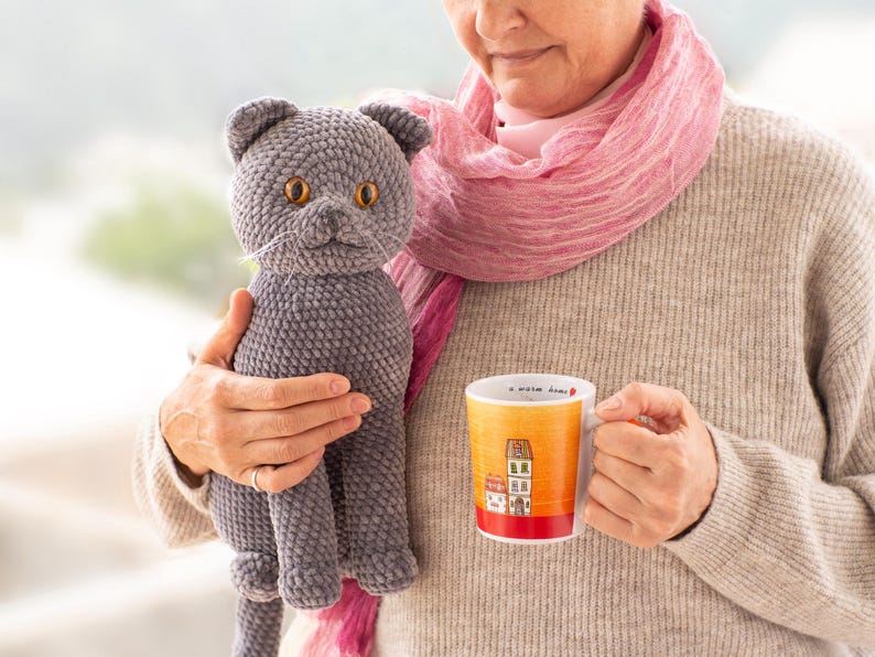 May include: A gray crocheted cat with orange eyes and a white whisker is held next to a mug. The mug has a yellow and orange gradient with a red base and features a house illustration. The cat is a plush toy.