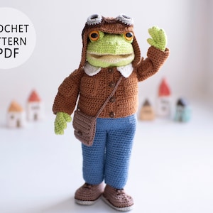 May include: A crocheted frog doll wearing a brown aviator jacket, goggles, and a matching hat. The frog has green skin, blue pants, and brown shoes. A small brown satchel hangs from its shoulder. The text "CROCHET PATTERN PDF" is in a white circle.