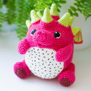 Op de afbeelding: Een roze en groene gehaakte draak speelgoed met een wit en zwart drakenfruit ontwerp. Het speelgoed heeft een klein, rond lichaam en een lange staart.