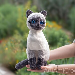 May include: A crocheted Siamese cat plush toy featuring a cream-colored body, dark gray ears, face, and paws, and bright blue plastic eyes. The cat is in a seated position with its tail curled forward. The text AniAmiCrochet is visible.