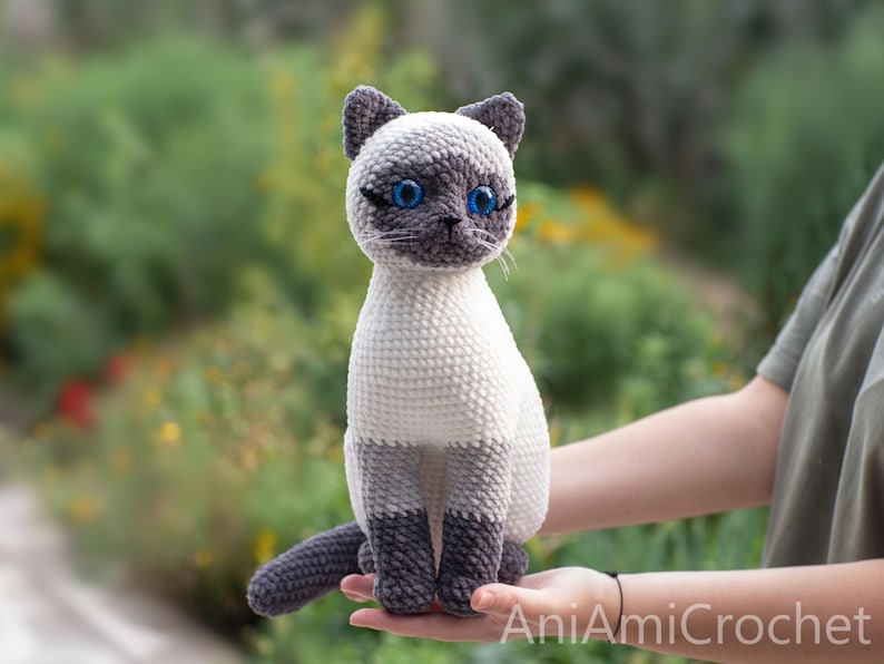 May include: A crocheted Siamese cat plush toy featuring a cream-colored body, dark gray ears, face, and paws, and bright blue plastic eyes. The cat is in a seated position with its tail curled forward. The text AniAmiCrochet is visible.