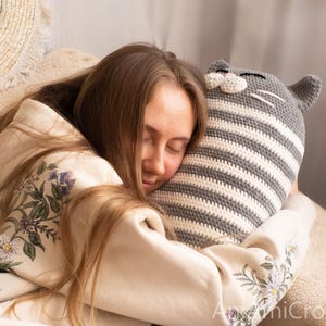 May include: A woman is sleeping with her head on a gray and white striped crocheted cat pillow. The pillow has a cute face with white whiskers.