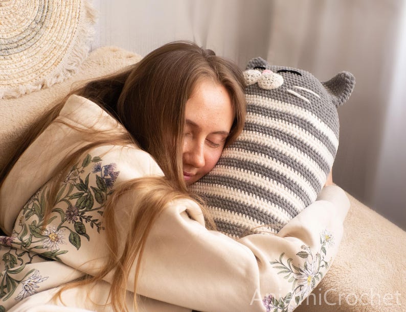 May include: A woman is sleeping with her head on a gray and white striped crocheted cat pillow. The pillow has a cute face with white whiskers.