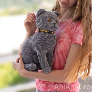 May include: A gray crocheted cat plush toy with a textured, velvety appearance. The cat has amber-colored eyes, a small stitched nose, and a yellow and black plaid collar featuring a small silver bell. The text AniAmiCrochet is visible.