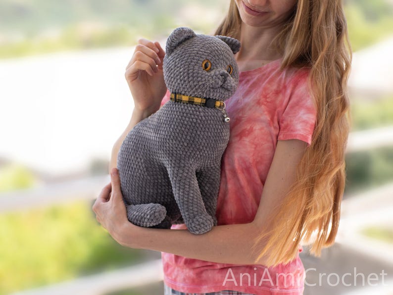 May include: A gray crocheted cat plush toy with a textured, velvety appearance. The cat has amber-colored eyes, a small stitched nose, and a yellow and black plaid collar featuring a small silver bell. The text AniAmiCrochet is visible.