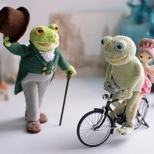 May include: A green crocheted frog wearing a brown top hat and a green jacket with a white collar and gold buttons, holding a brown walking stick. A second green crocheted frog is riding a black bicycle with a pink crocheted fairy sitting on the back.