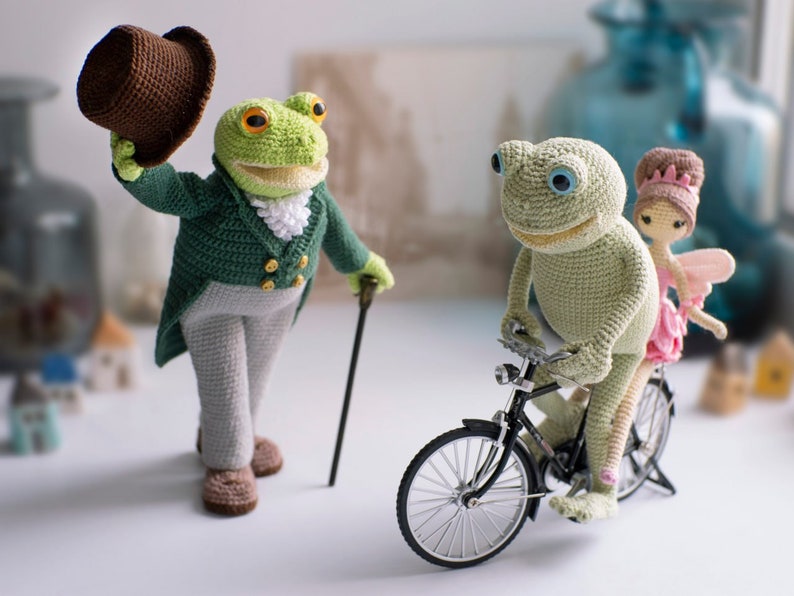 May include: A green crocheted frog wearing a brown top hat and a green jacket with a white collar and gold buttons, holding a brown walking stick. A second green crocheted frog is riding a black bicycle with a pink crocheted fairy sitting on the back.
