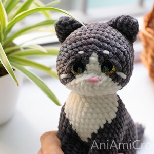 May include: A handmade crochet cat plush toy featuring dark gray and white yarn, large glossy black eyes with gold glitter accents, and a small pink nose. The text AniAmiCrochet is visible in the bottom right corner of the image.