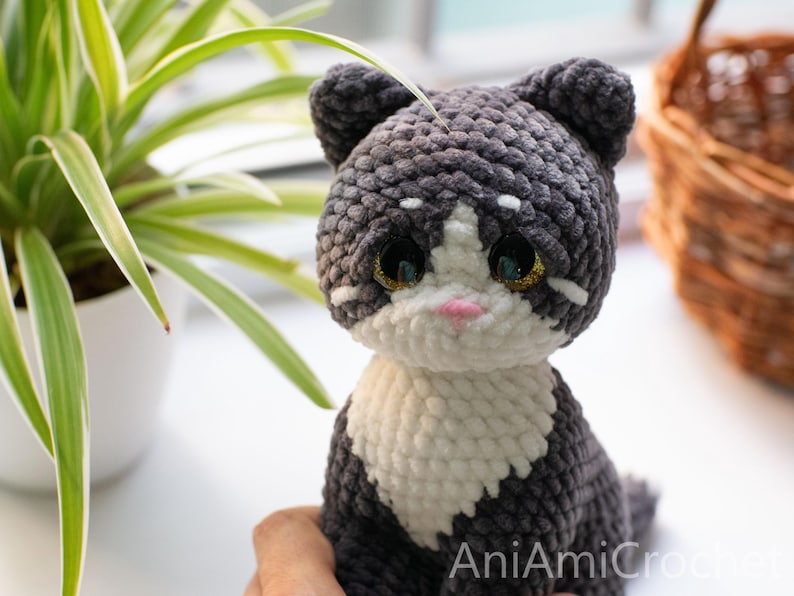 May include: A handmade crochet cat plush toy featuring dark gray and white yarn, large glossy black eyes with gold glitter accents, and a small pink nose. The text AniAmiCrochet is visible in the bottom right corner of the image.