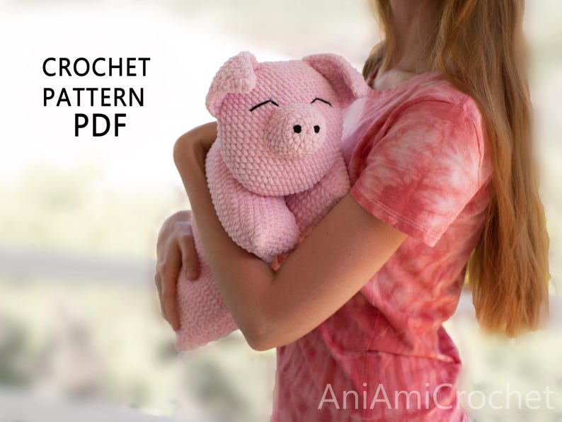 K&ouml;nnte beinhalten: Ein geh&auml;keltes rosa Schwein-Stofftier mit strukturierter Garnoberfl&auml;che, Schlappohren und einer Schnauze mit zwei schwarzen Nasenl&ouml;chern. Der Text CROCHET PATTERN PDF ist oben links und AniAmiCrochet unten rechts zu sehen.