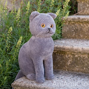 May include: A handmade, gray crocheted cat figurine with yellow eyes and a small black nose. The cat is sitting on a stone step, with green foliage in the background. The cat has a textured, plush appearance.