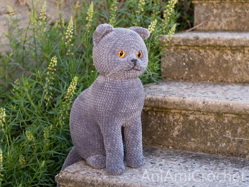 May include: A handmade, gray crocheted cat figurine with yellow eyes and a small black nose. The cat is sitting on a stone step, with green foliage in the background. The cat has a textured, plush appearance.