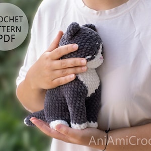 May include: A crochet pattern PDF for a plush cat toy. The cat is made of dark gray and white yarn with a soft, textured finish. It features a white chest, white paws, and a small pink nose. The text CROCHET PATTERN PDF is visible in the upper left corner.