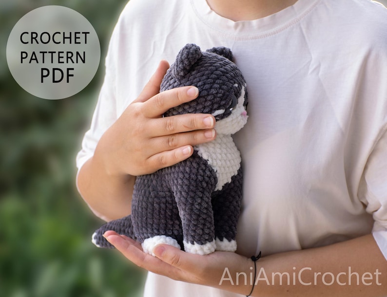 May include: A crochet pattern PDF for a plush cat toy. The cat is made of dark gray and white yarn with a soft, textured finish. It features a white chest, white paws, and a small pink nose. The text CROCHET PATTERN PDF is visible in the upper left corner.