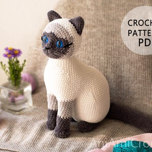 May include: A crocheted Siamese cat toy with a cream-coloured body, dark brown ears, face, paws, and tail, and bright blue eyes. The image includes a circular white graphic with the text CROCHET PATTERN PDF and the watermark AniAmiCrochet.