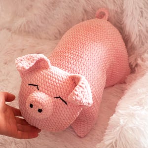 K&ouml;nnte beinhalten: Ein hellrosa geh&auml;keltes Schweinchen-Pl&uuml;schtier mit einer abgerundeten Schnauze, zwei schwarzen gestickten Nasenl&ouml;chern, zwei dreieckigen Ohren und geschlossenen Augen, die durch einfache schwarze horizontale Stiche dargestellt sind. Es hat einen kleinen Ringelschwanz.